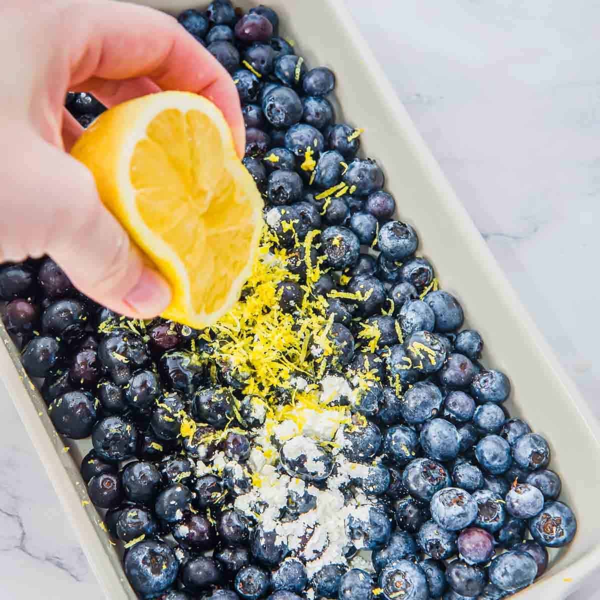 A hand squeezes a lemon half over a baking dish filled with fresh blueberries, lemon zest, and a sprinkle of flour.