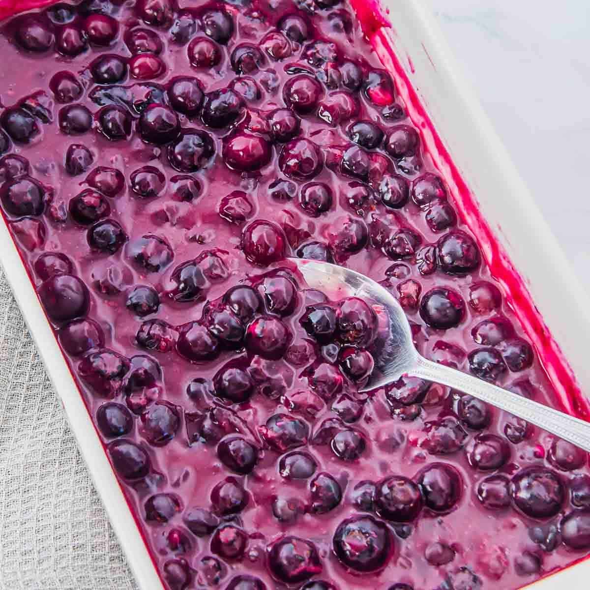 A rectangular white dish filled with glossy baked blueberries, with a silver spoon resting inside, surrounded by a textured gray cloth on a white surface.