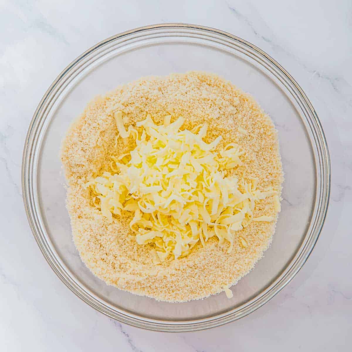 A glass bowl containing a mixture of flour and grated butter sits on a white marble surface.