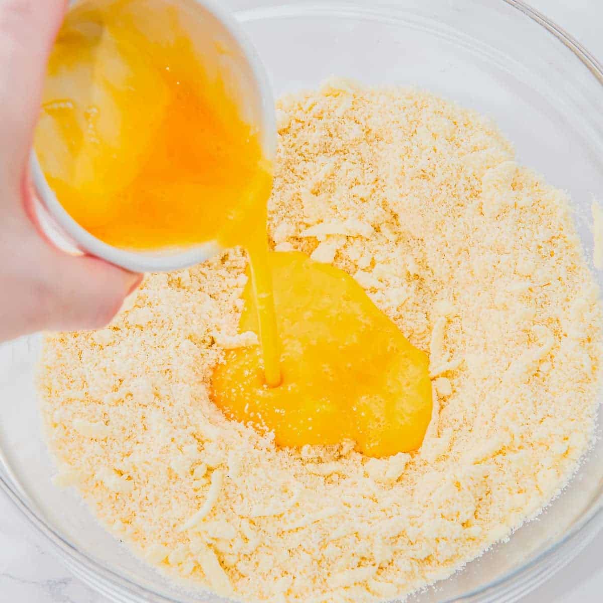 A hand pours beaten eggs from a small bowl into a large glass bowl filled with a crumbly flour mixture, preparing to mix ingredients for baking.