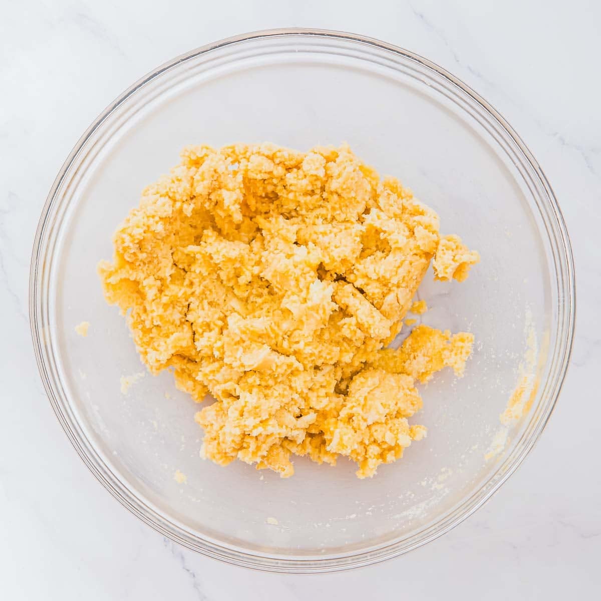 A clear glass bowl containing a yellow, crumbly cobbler dough mixture sits on a white marble surface.
