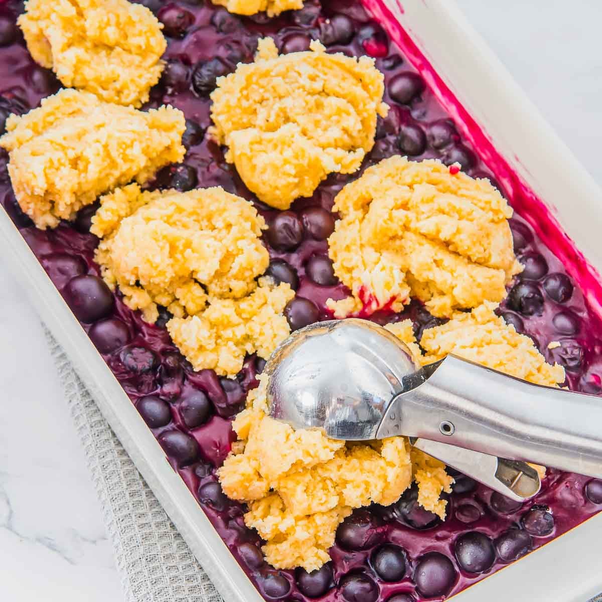 A baking dish filled with blueberries topped with scoops of yellow biscuit dough, placed with a metal ice cream scoop, ready to be baked.