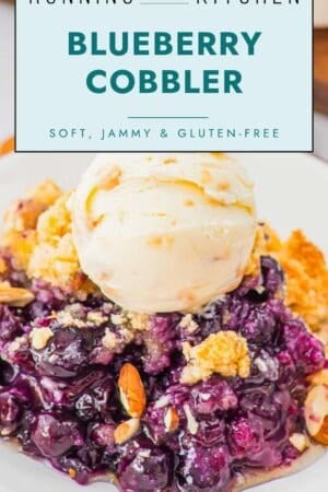 A close-up of blueberry cobbler topped with a scoop of vanilla ice cream. The cobbler filling is jammy with whole blueberries and a golden crumb topping. Text above reads "Blueberry Cobbler - Soft, Jammy & Gluten-Free.