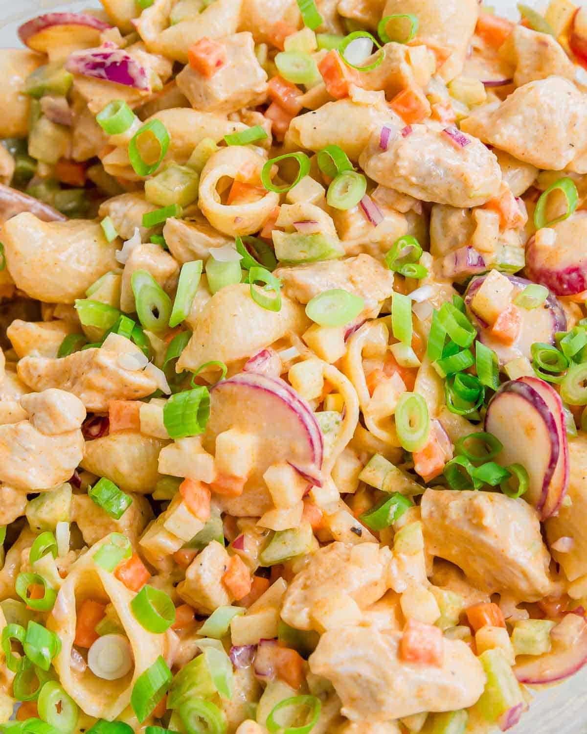 A close-up of a creamy buffalo chicken salad with cooked pasta shells, diced vegetables, radish slices, and chopped green onions sprinkled on top. The salad appears fresh and colorful.