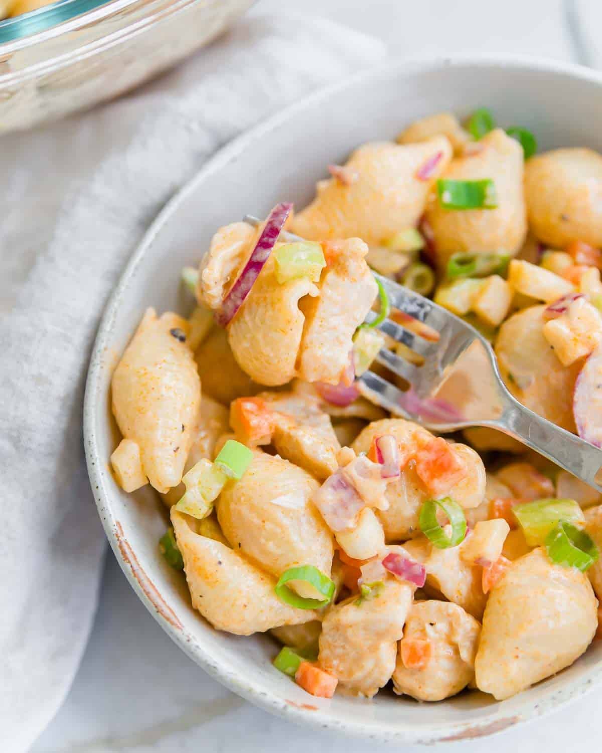 A close-up of a bowl of creamy buffalo chicken pasta salad with shell pasta, diced vegetables, green onions, and a fork picking up a bite. The salad is served in a light-colored bowl on a white surface.