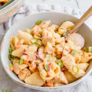 A bowl of buffalo chicken pasta salad with small shell pasta, diced chicken, green onions, red onions, carrots, and bell peppers, served with a fork on a light cloth background.