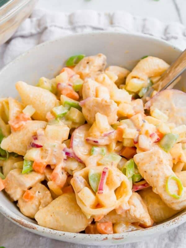 A bowl of buffalo chicken pasta salad with small shell pasta, diced chicken, green onions, red onions, carrots, and bell peppers, served with a fork on a light cloth background.