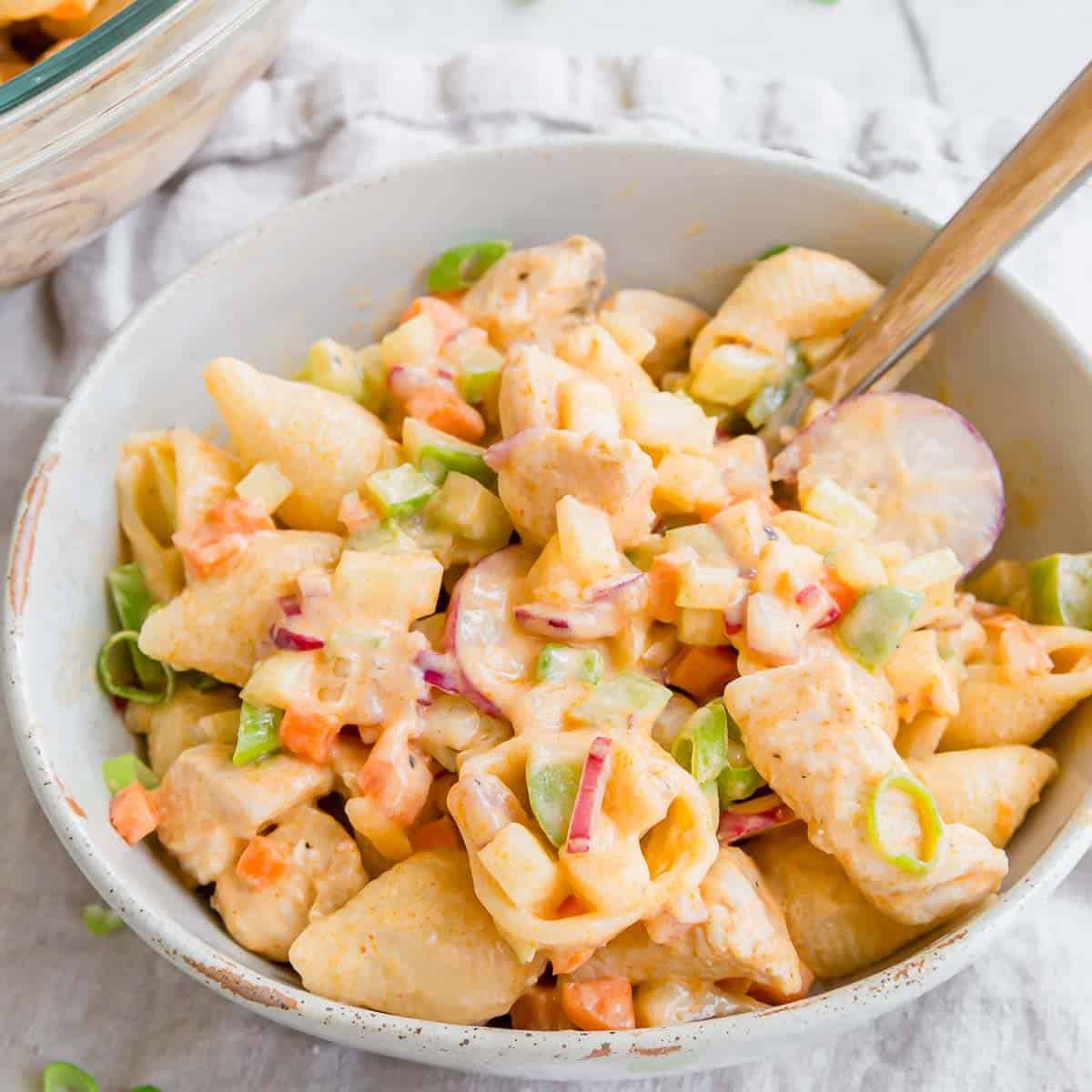 A bowl of buffalo chicken pasta salad with small shell pasta, diced chicken, green onions, red onions, carrots, and bell peppers, served with a fork on a light cloth background.