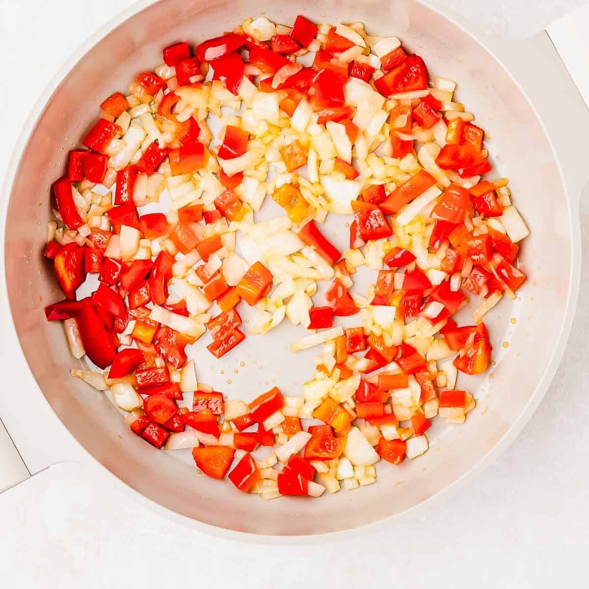 Diced red bell peppers and onions are being sautéed in a white skillet.