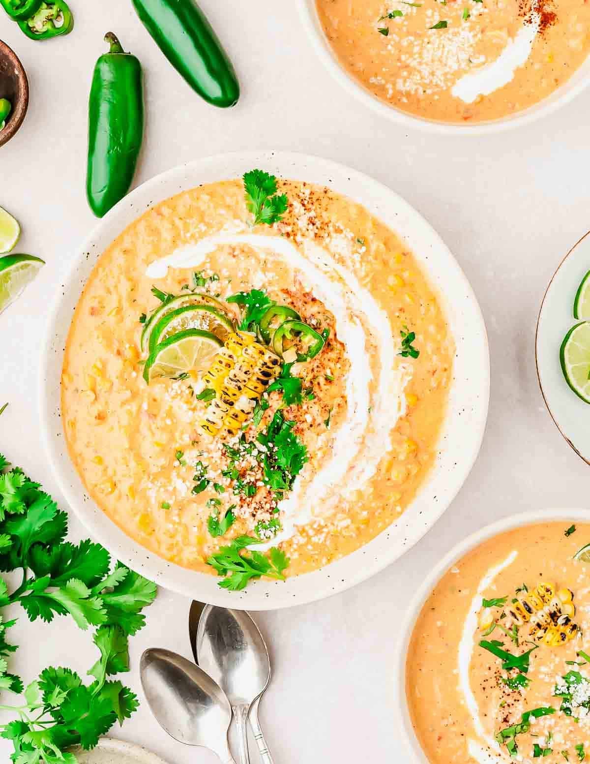 A bowl of Mexican street corn soup garnished with grilled corn, lime slices, fresh cilantro, sliced jalapeño, and a swirl of cream, surrounded by more soup bowls, jalapeños, lime wedges, cilantro, and two silver spoons.