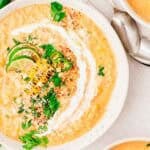 Creamy corn soup garnished with grilled corn, lime slices, jalapeño, fresh cilantro, crumbled cheese, and a swirl of cream, served in a white bowl with a spoon nearby.