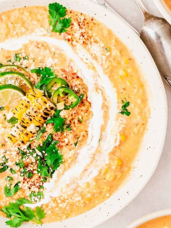 Creamy corn soup garnished with grilled corn, lime slices, jalapeño, fresh cilantro, crumbled cheese, and a swirl of cream, served in a white bowl with a spoon nearby.