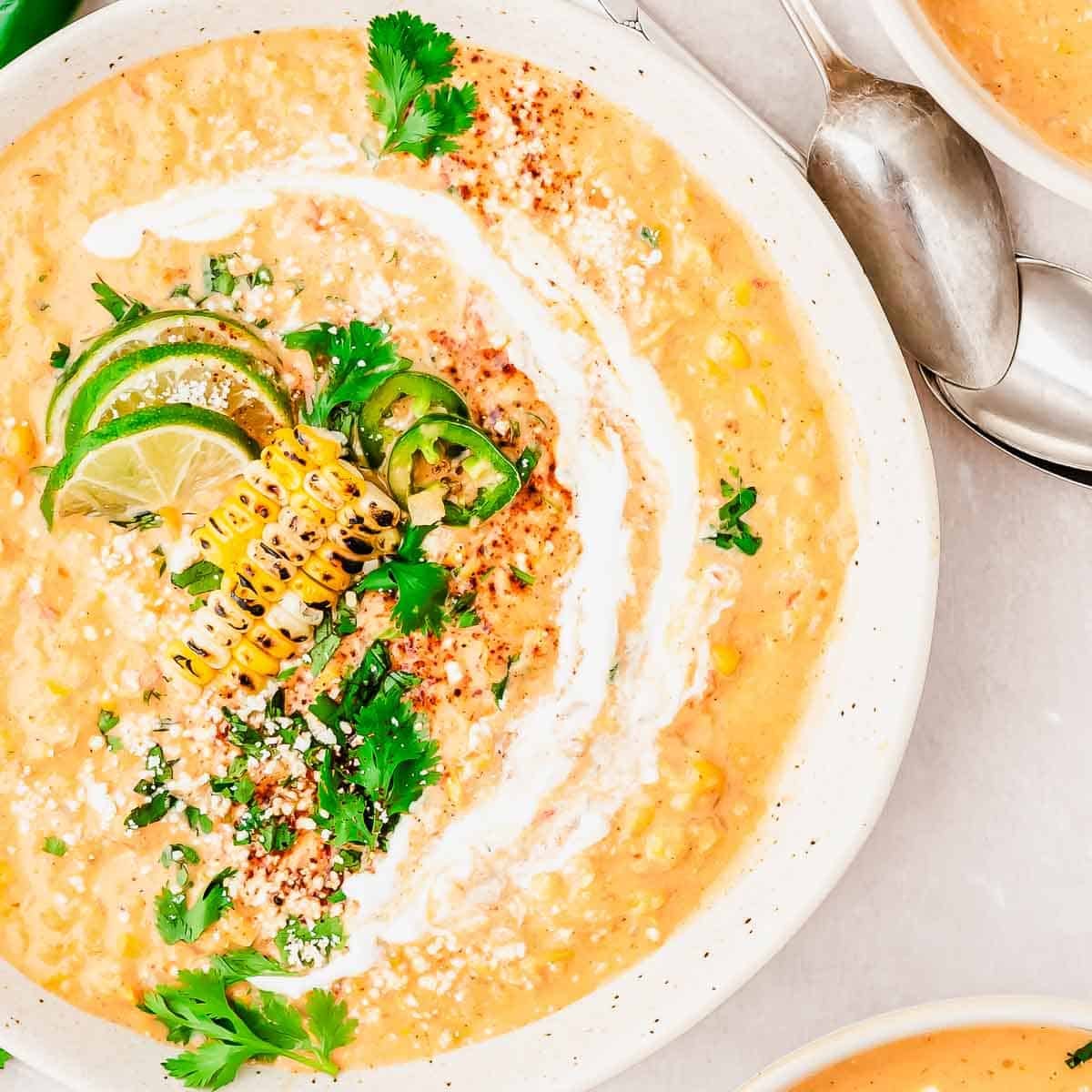 Creamy corn soup garnished with grilled corn, lime slices, jalapeño, fresh cilantro, crumbled cheese, and a swirl of cream, served in a white bowl with a spoon nearby.