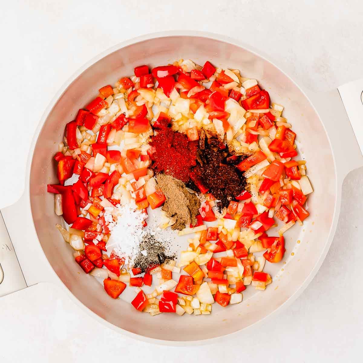 A white pot with chopped red bell peppers, onions, and piles of spices including black pepper, salt, paprika, and cumin, arranged before mixing.