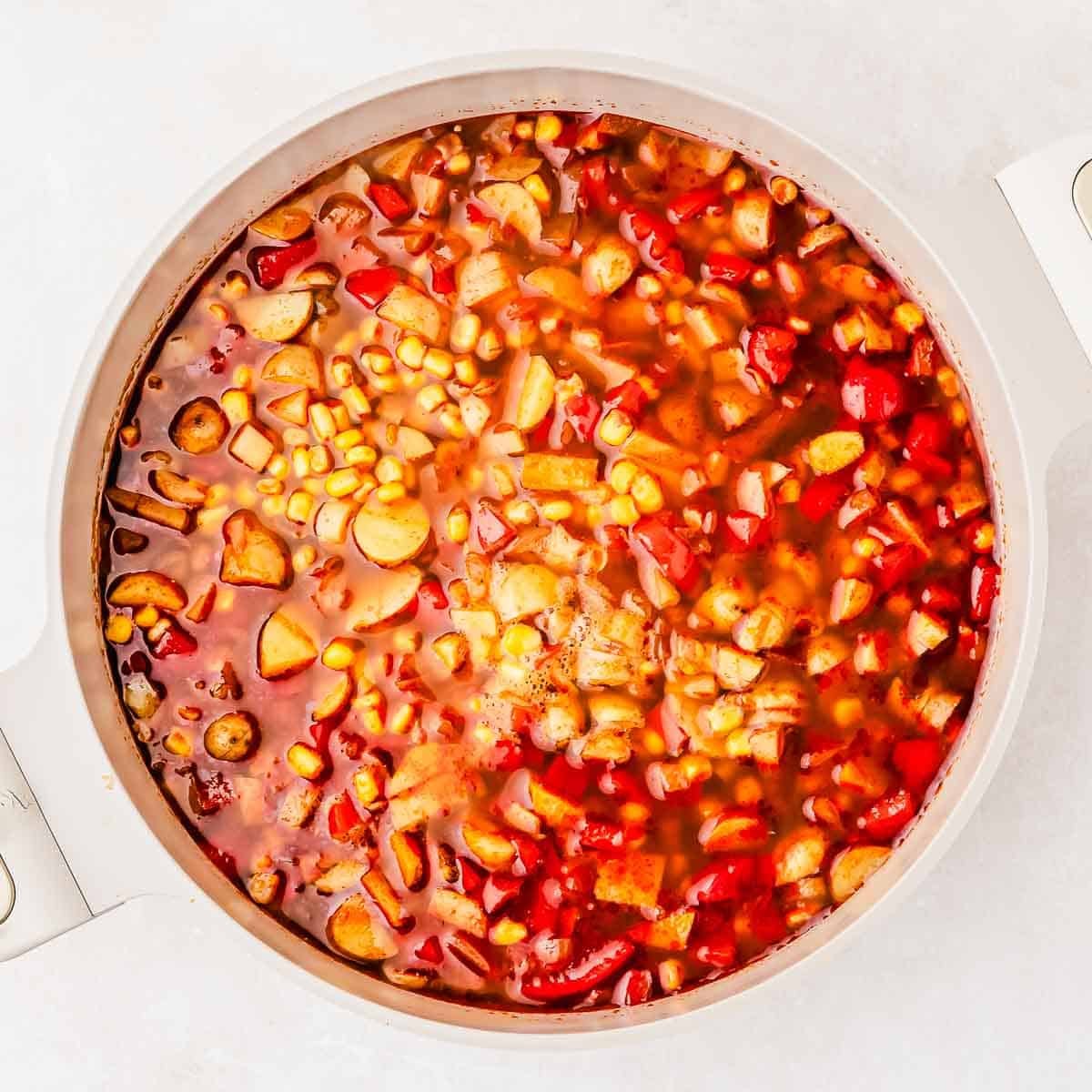 A pot filled with a colorful soup, featuring visible corn, red bell peppers, potatoes, and a rich red broth.