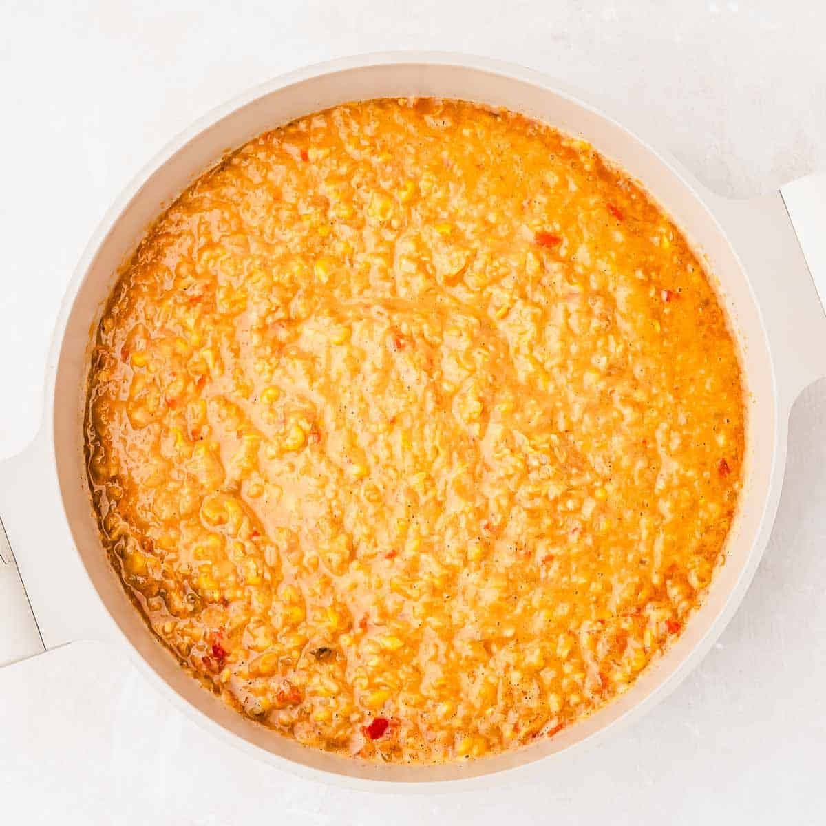 A round white pot filled with creamy orange corn soup featuring visible small bits of vegetables, sits on a light surface.