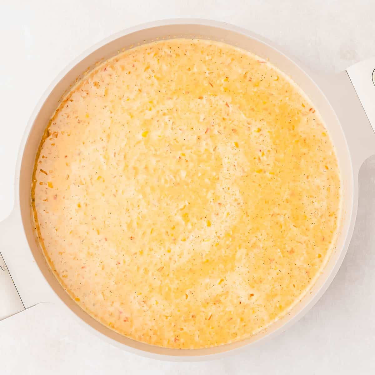 A creamy, yellow corn soup simmering in a light-colored pot, viewed from above on a light surface. Small bits of herbs and ingredients are visible throughout the soup.