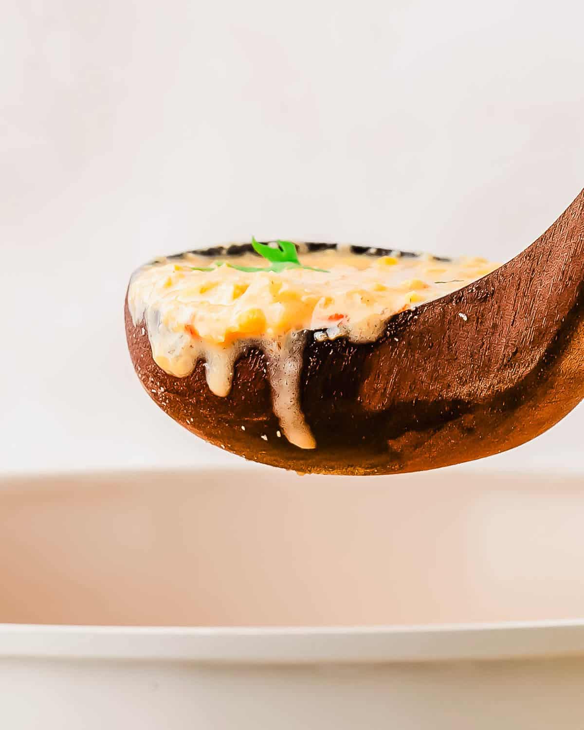 A wooden ladle holds creamy soup with corn and herbs, dripping slightly over the edge, above a white bowl.