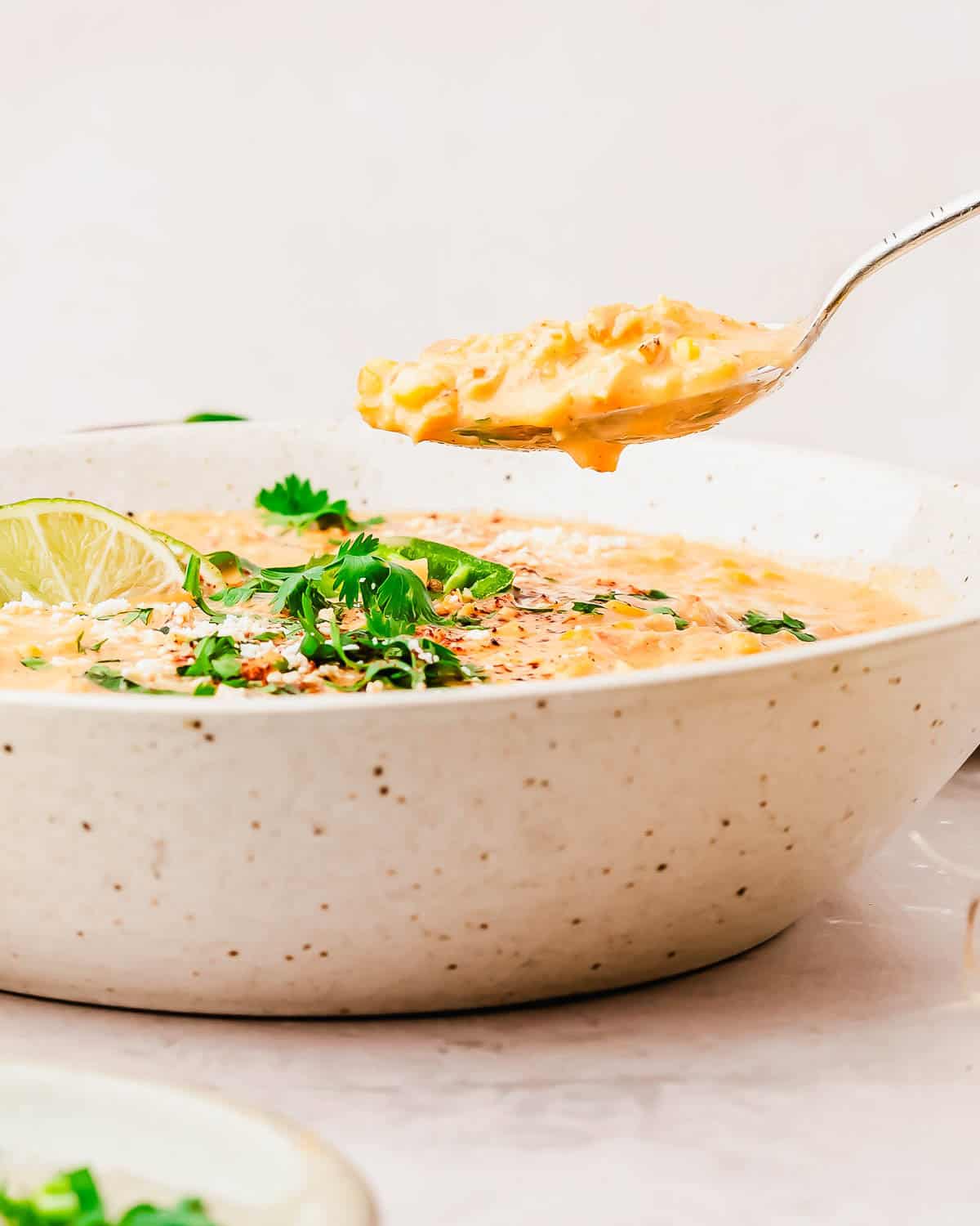 A close-up of a bowl of creamy street corn soup garnished with cilantro, lime, and sesame seeds, with a spoon lifting a bite from the bowl.