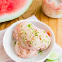 Three scoops of watermelon ice cream garnished with lime zest in a white bowl, with lime slices and a jar of ice cream nearby on a wooden surface. A watermelon is visible in the background.
