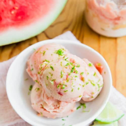 Three scoops of watermelon ice cream garnished with lime zest in a white bowl, with lime slices and a jar of ice cream nearby on a wooden surface. A watermelon is visible in the background.