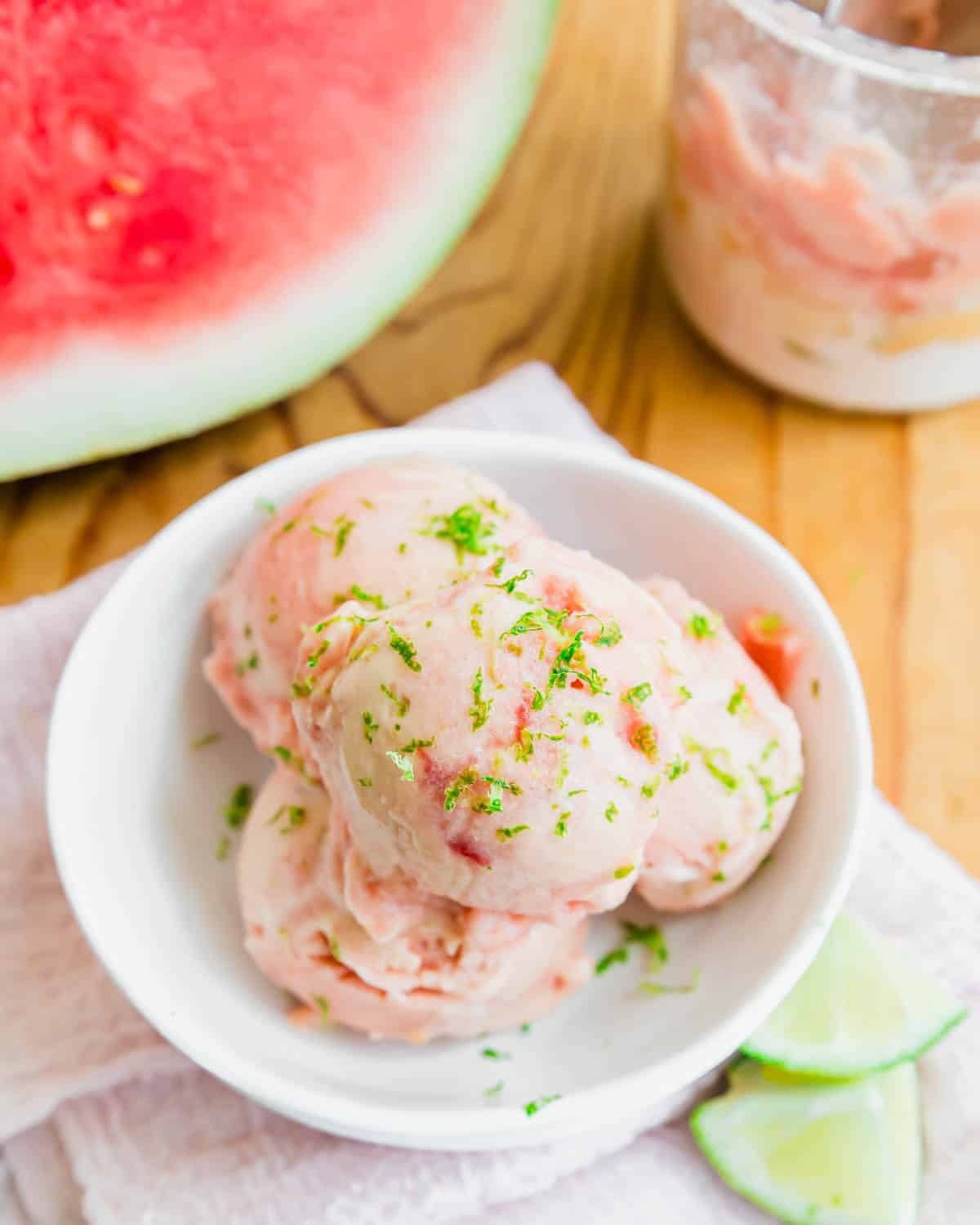 Three scoops of watermelon ice cream garnished with lime zest in a white bowl, with lime slices and a jar of ice cream nearby on a wooden surface. A watermelon is visible in the background.