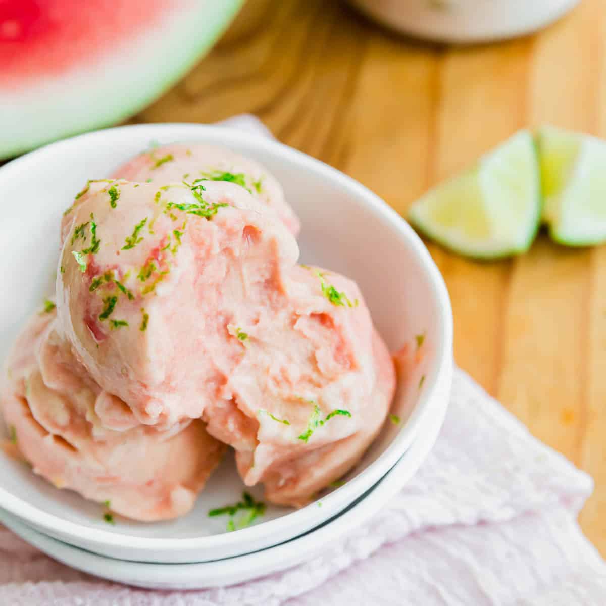 Two scoops of pink watermelon ice cream in a white bowl, garnished with lime zest, on a white napkin. A partially visible watermelon and lime wedges are in the background on a wooden surface.