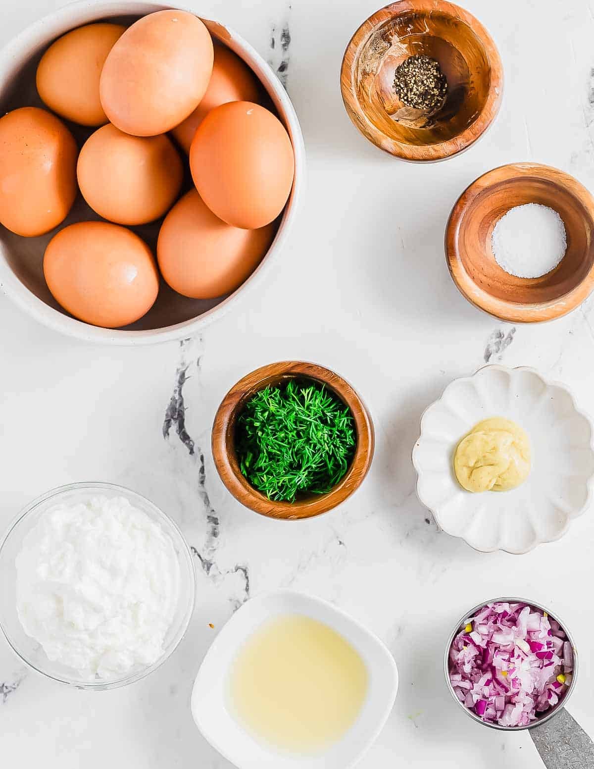 Eggs in a bowl, with small bowls of black pepper, salt, chopped dill, mustard, chopped red onion, yogurt, and a dish of vinegar arranged on a white marble surface.