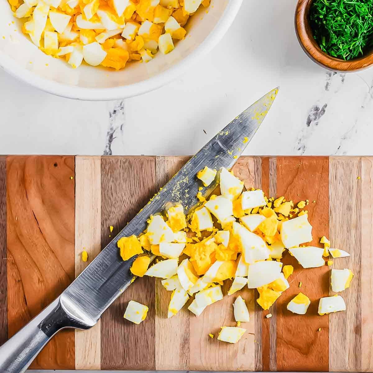 Chopped hard-boiled eggs and a knife on a striped wooden cutting board, with a bowl of chopped eggs and a small bowl of herbs nearby on a marble countertop.