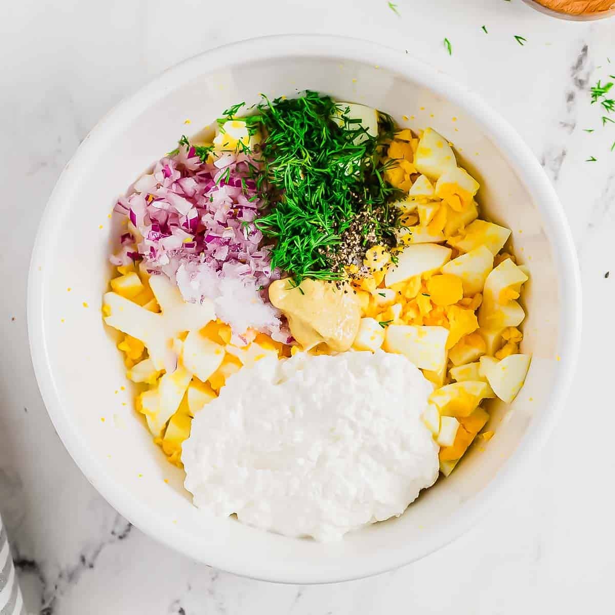 A white bowl containing chopped hard-boiled eggs, red onion, fresh dill, black pepper, mustard, and a dollop of cottage cheese on a white marble surface.