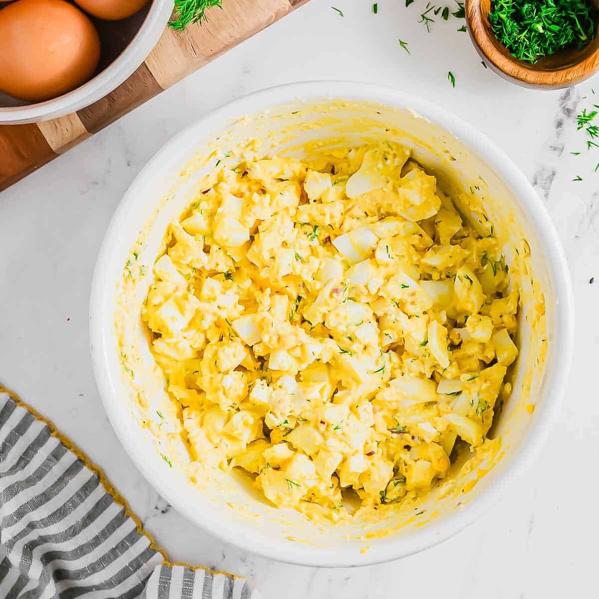 A white bowl filled with creamy cottage cheese egg salad sits on a marble countertop, surrounded by a bowl of eggs, a small bowl of herbs, and a striped cloth napkin.