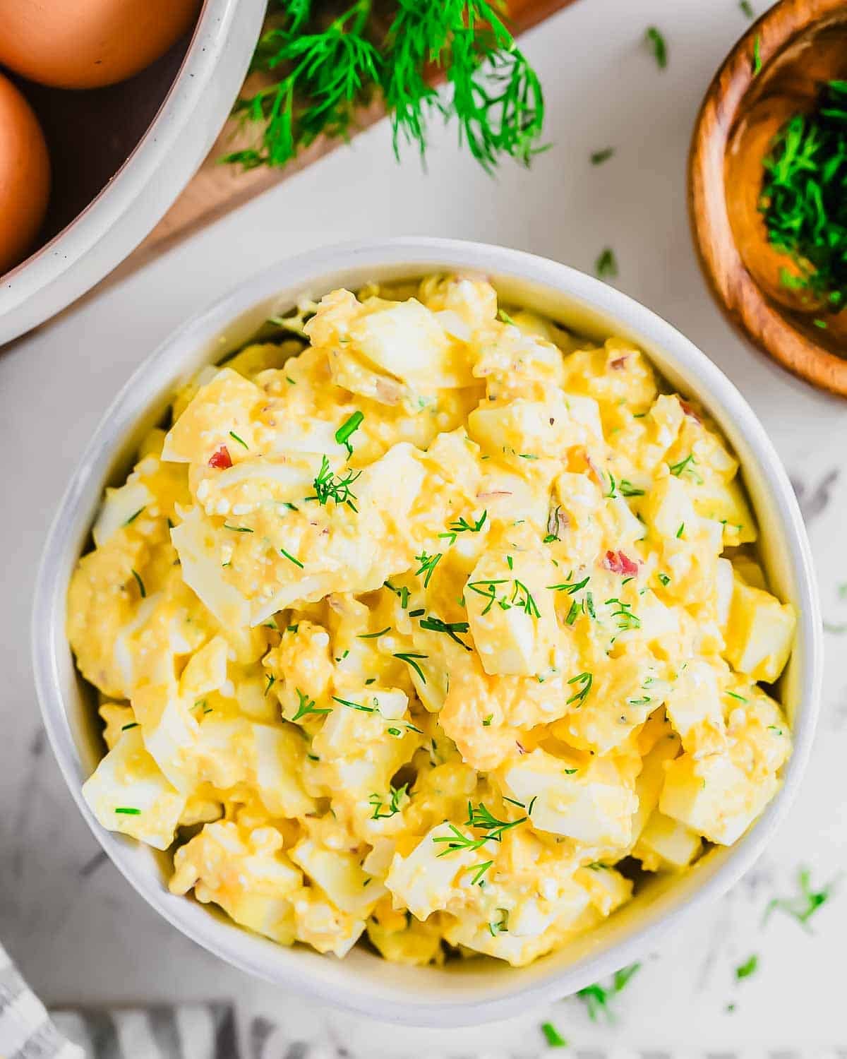 A bowl of creamy egg salad garnished with fresh chopped dill, surrounded by ingredients like eggs and dill sprigs on a white surface.