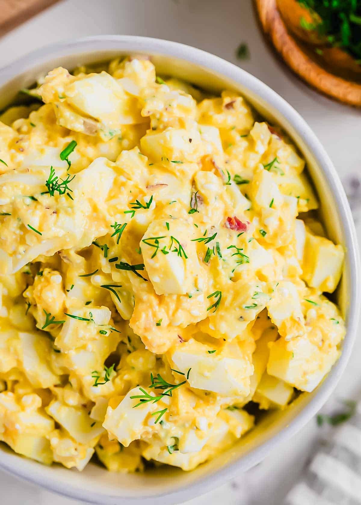 A close-up of a bowl filled with cottage cheese egg salad garnished with fresh dill. The salad contains chopped eggs, mayonnaise, and herbs, and is placed on a light-colored surface.