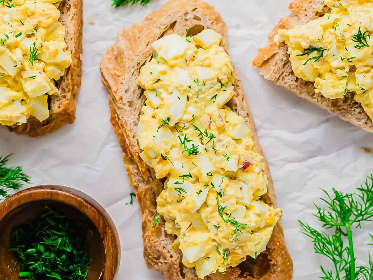 Open-faced egg salad sandwiches on crusty bread, garnished with fresh dill, displayed on parchment paper with a small wooden bowl of chopped herbs nearby.