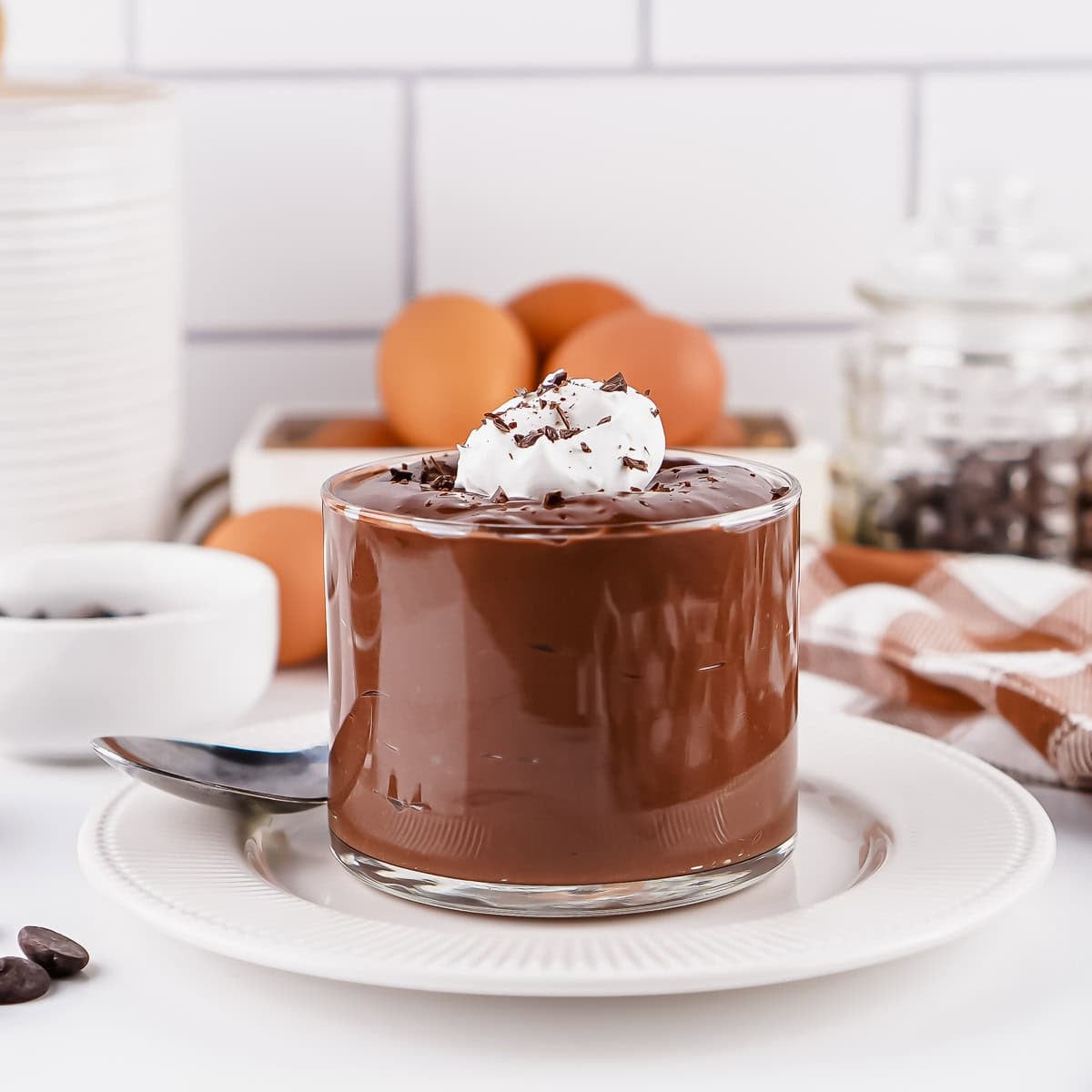 A glass of chocolate pudding topped with whipped cream and chocolate shavings sits on a white plate with a spoon, eggs, and coffee beans in the background.