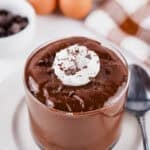 A glass cup filled with chocolate egg pudding, topped with whipped cream and chocolate shavings. A spoon, two eggs, a bowl of chocolate chips, and a checkered napkin are in the background.