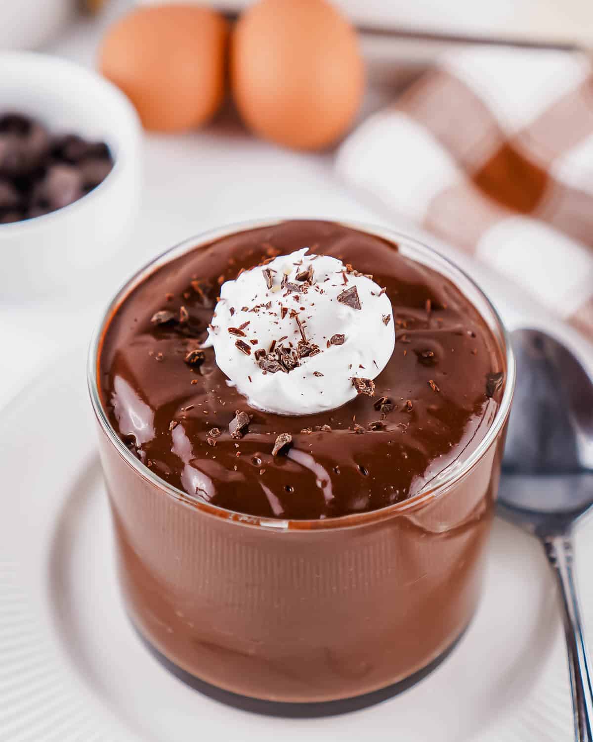 A glass cup filled with chocolate egg pudding, topped with whipped cream and chocolate shavings. A spoon, two eggs, a bowl of chocolate chips, and a checkered napkin are in the background.