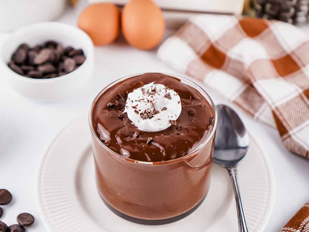 A glass cup of hard-boiled egg chocolate pudding topped with whipped cream and chocolate shavings sits on a white plate next to a spoon, with eggs and chocolate chips in the background.