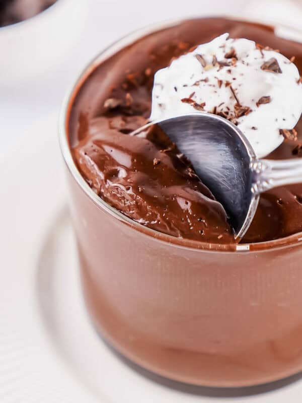 A glass of chocolate egg pudding topped with whipped cream and chocolate shavings, with a spoon scooping some from the top, on a white plate.