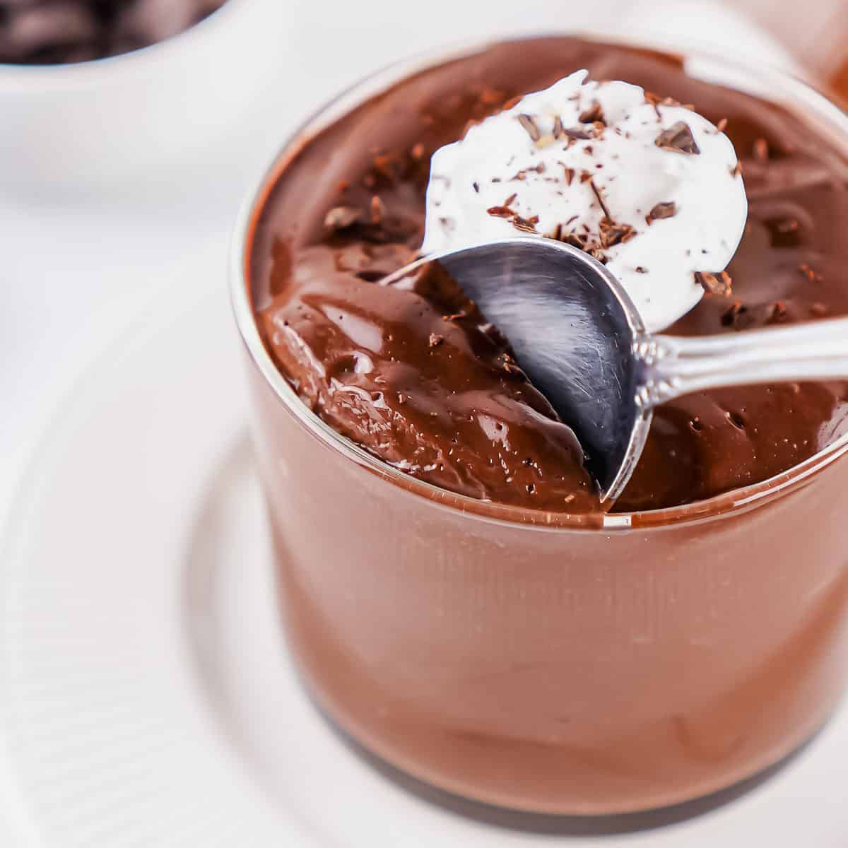 A glass of chocolate egg pudding topped with whipped cream and chocolate shavings, with a spoon scooping some from the top, on a white plate.