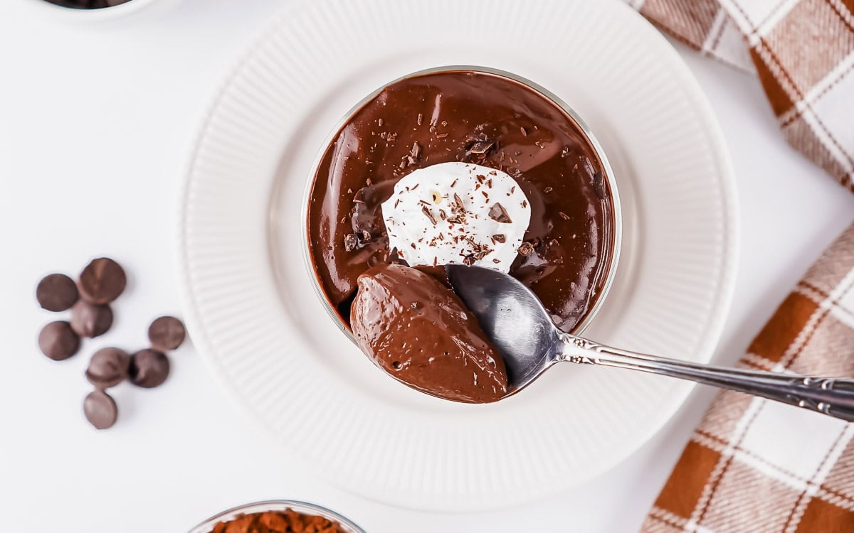 A bowl of chocolate pudding topped with whipped cream and chocolate shavings, with a spoon scooping some out. Chocolate chips and a checkered napkin are nearby.