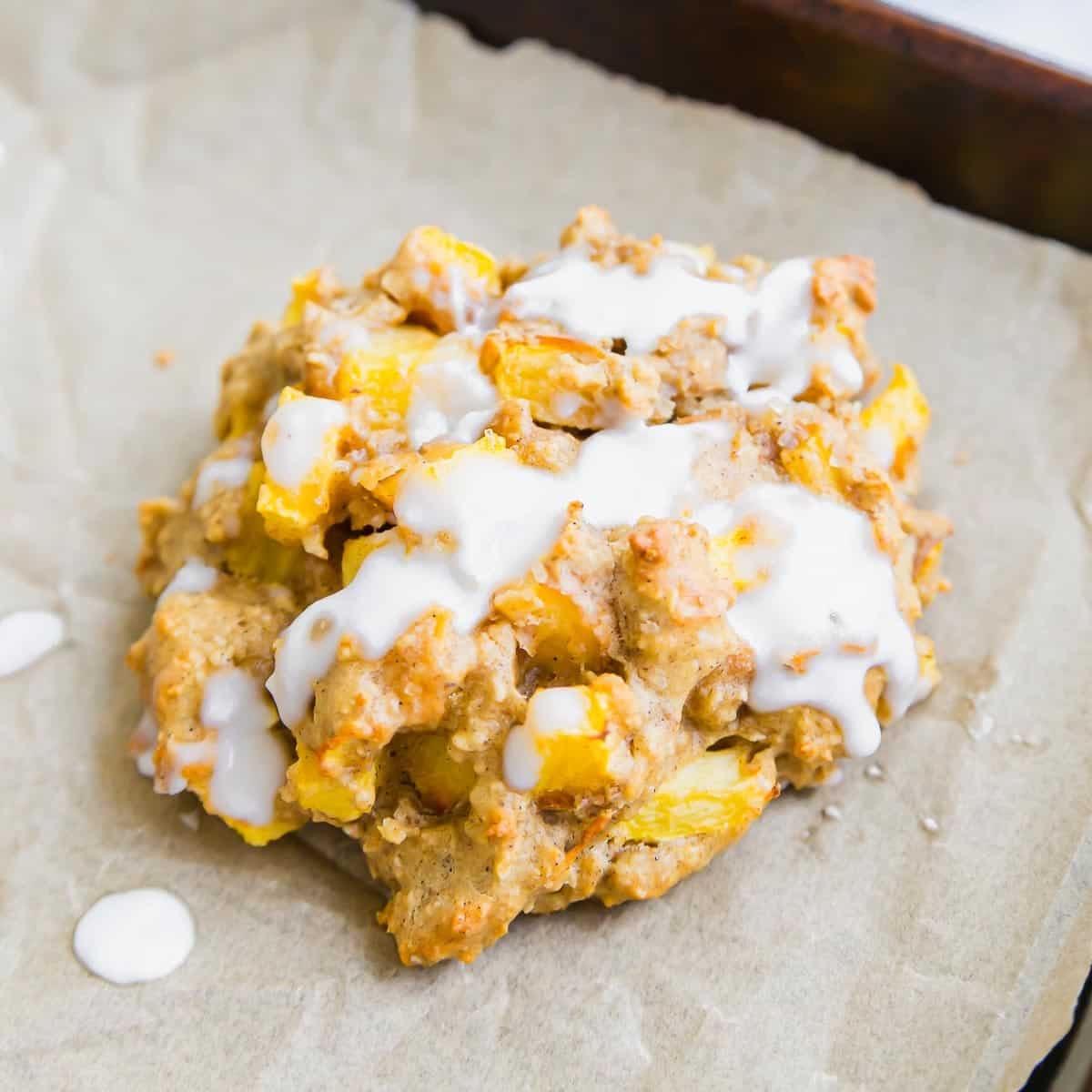 A chunky baked fritter with visible yellow fruit pieces and a drizzle of white icing sits on a piece of parchment paper.