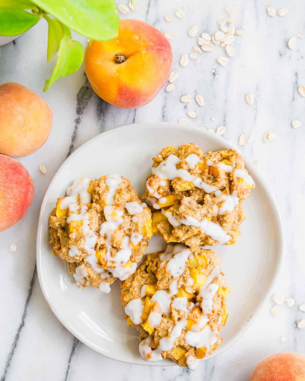 Three peach fritters with white icing on a round white plate, surrounded by fresh peaches and scattered oats on a marble surface. A green plant is partially visible in the upper left corner.