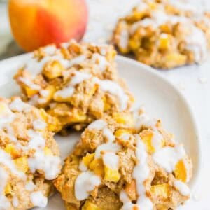 Three baked peach fritters with white icing drizzle are on a white plate. A fresh peach and more fritters are blurred in the background. The cookies have chunks of peach and a soft, rustic texture.