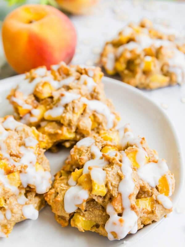 Three baked peach fritters with white icing drizzle are on a white plate. A fresh peach and more fritters are blurred in the background. The cookies have chunks of peach and a soft, rustic texture.