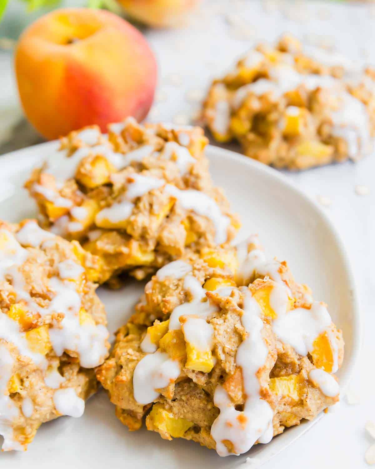 Three baked peach fritters with white icing drizzle are on a white plate. A fresh peach and more fritters are blurred in the background. The cookies have chunks of peach and a soft, rustic texture.