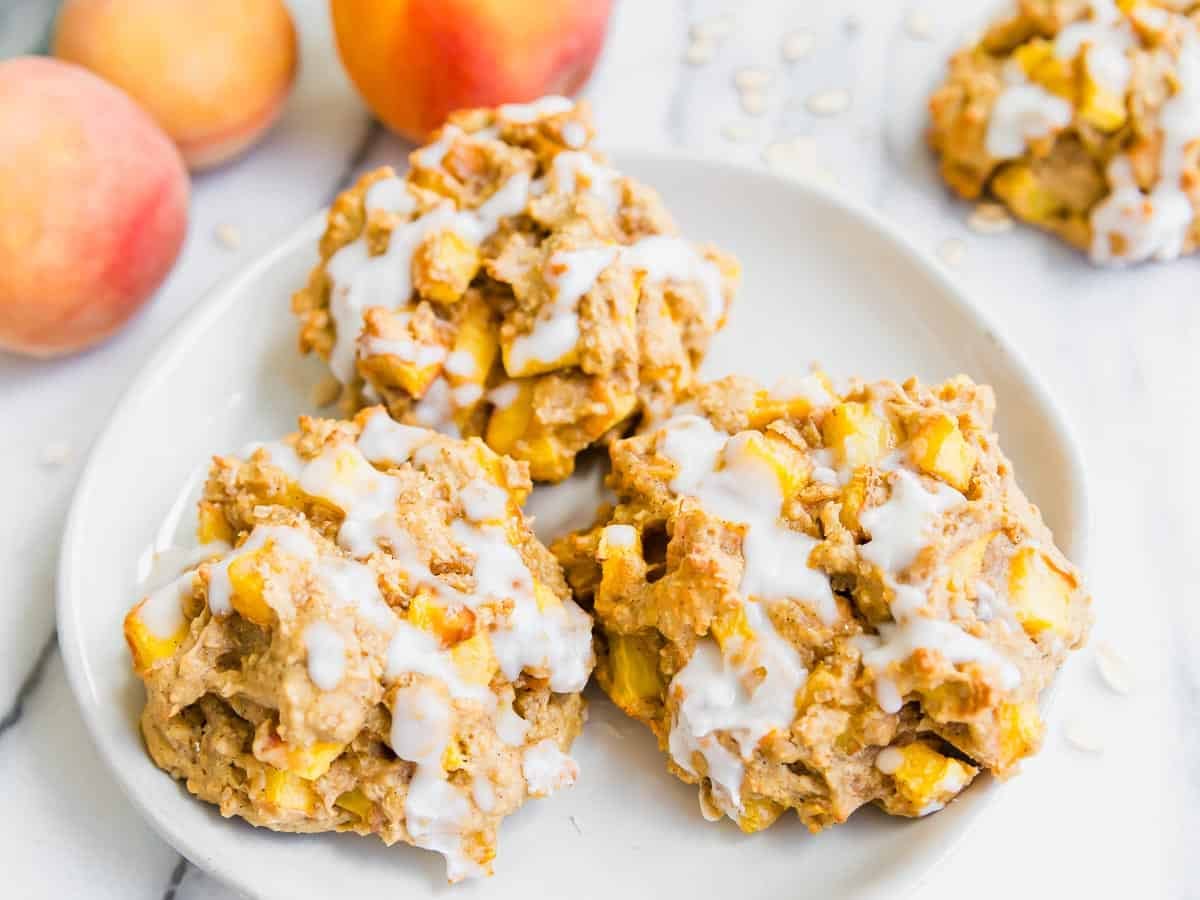 Three gluten-free peach fritters with white icing drizzle are on a white plate. Fresh peaches and more fritters are blurred in the background. The fritters are chunky and filled with visible peach pieces.