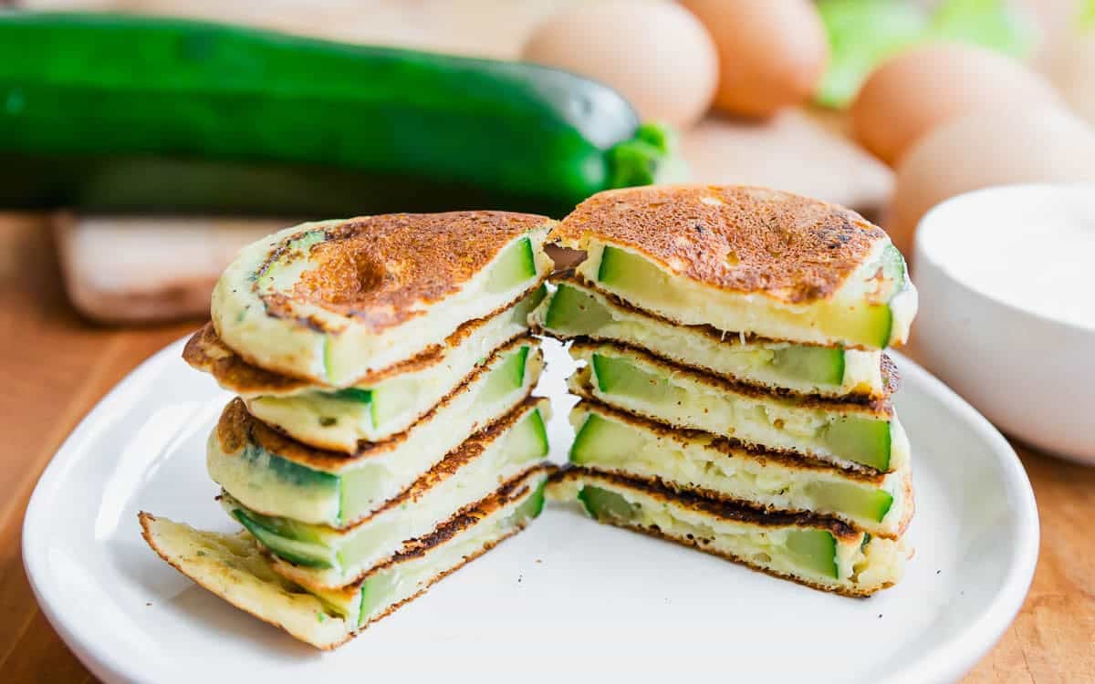 A stack of round, sliced zucchini pancakes cut in half, showing green zucchini pieces inside. Eggs, a zucchini, and a bowl are blurred in the background.