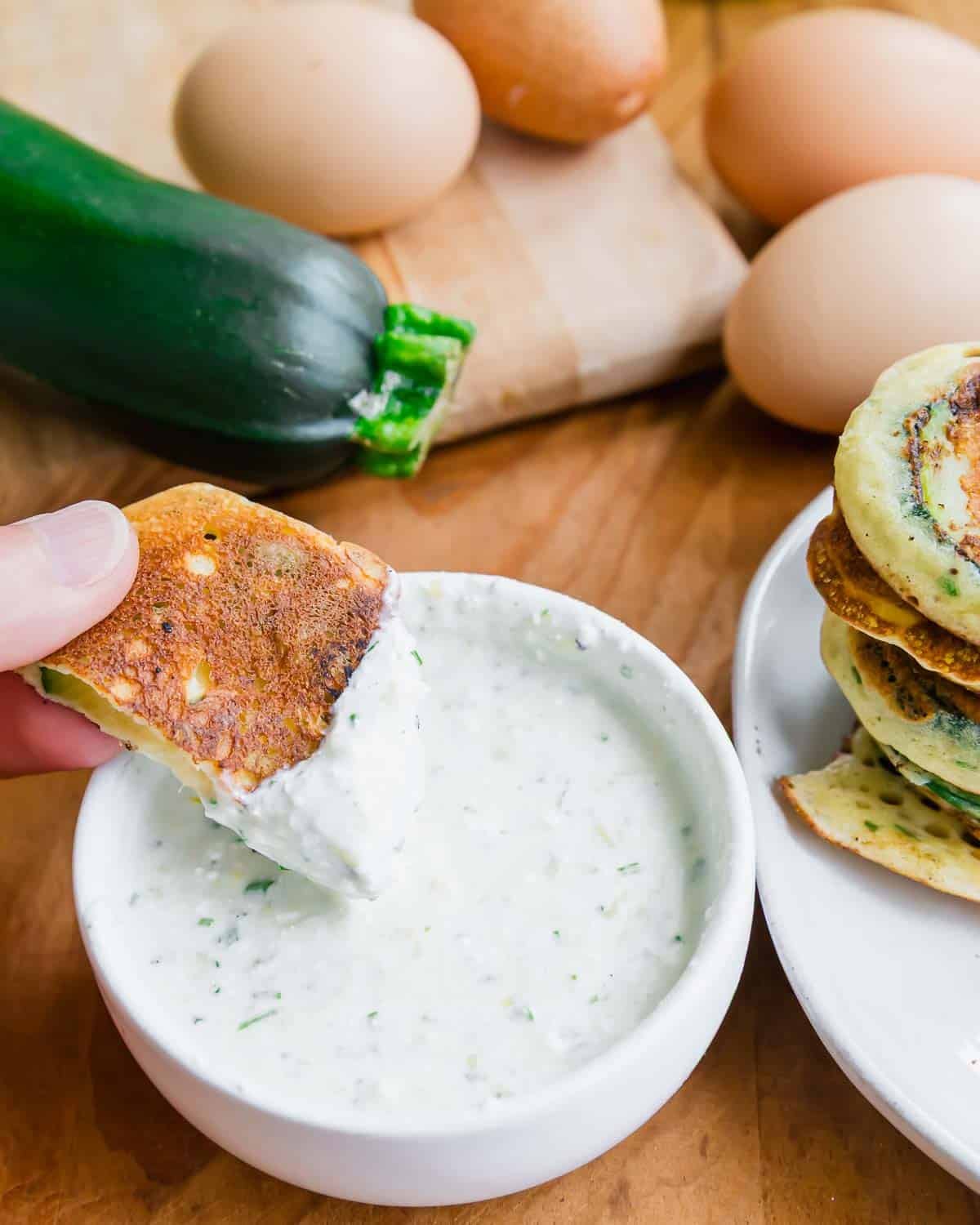 A hand dips a golden zucchini pancake into a bowl of creamy feta dip, with eggs and a zucchini in the background on a wooden surface.