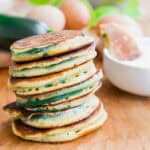 A stack of golden-brown zucchini pancakes sits on a wooden surface, with eggs, a zucchini, and a white bowl in the background.